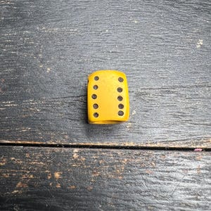 May include: A yellow die with black dots, showing the numbers 1, 2, 3, 4, 5, and 6. The die is rectangular and has rounded edges. The background is a dark, weathered wooden surface.