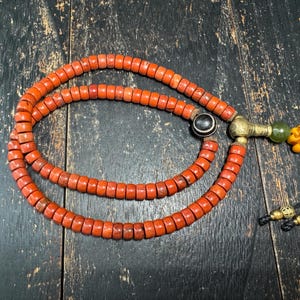 May include: A strand of cylindrical orange beads arranged in a circular pattern. The necklace features a single black and white striped bead, a metallic connector, a green bead, and a yellow knot ornament with two black cords ending in gold beads.