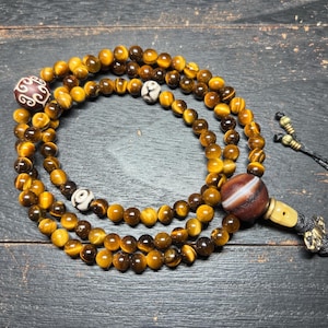 May include: A layered mala bead necklace featuring tiger's eye beads in shades of brown and gold. The necklace includes decorative beads with intricate patterns and a tassel. The beads are strung on a black cord.