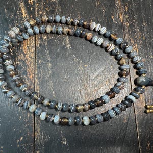 Puede incluir: Un collar largo de cuentas circulares de piedra gris y marrón con bandas, intercaladas con pequeños espaciadores metálicos. Incluye una cuenta central gris lisa, un dije metálico con forma de calavera y dos terminales metálicos en un cordón negro.