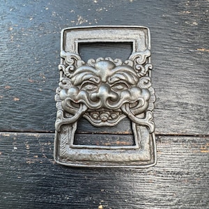 May include: A rectangular, silver-colored metal bottle opener with a detailed, ornate face design. The face has a prominent nose, smiling mouth, and elaborate decorative elements. The opener is set against a dark, textured wooden surface.
