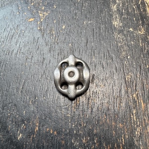 May include: A small, antique-style metal object with a circular design and four symmetrical openings. The object has a central hole and a textured, aged appearance, suggesting it is a vintage hardware component.