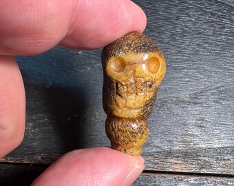 May include: A small, carved skull pendant in shades of tan and brown. The skull features detailed eye sockets, a nose, and teeth. The pendant is held between two fingers, showcasing its size and intricate design.
