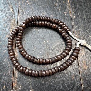 Op de afbeelding: Een streng donkerbruine gebedskralen met een witte draad en knoop. De kralen zijn rond en lijken gemaakt van hout of een vergelijkbaar materiaal. De kralen liggen op een donker houten oppervlak.