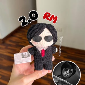 May include: A crochet doll with black hair, wearing black sunglasses, a black suit, and a red tie. The doll holds a small white rectangular card with printed text. The text 2.0 RM is visible above the doll, and a circular inset shows a person.