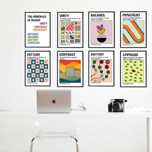 May include: Eight framed art prints on a white wall, each with a black border. The prints feature colorful abstract designs and text including "Unity," "Balance," and "Movement." A laptop, camera, and chair are in the foreground.