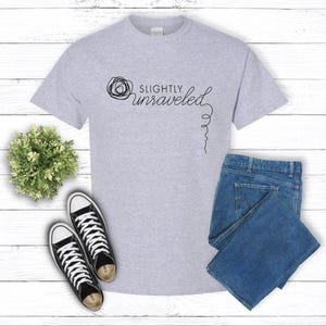 May include: A light gray t-shirt with the text "Slightly Unraveled" in black script, paired with blue denim jeans and black and white sneakers. A small green plant is also in the image.
