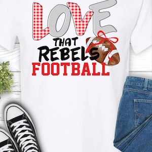 Puede incluir: Camiseta blanca con el texto "LOVE THAT REBELS FOOTBALL" en rojo, gris y negro. La palabra "LOVE" tiene un patrón de cuadros rojos y blancos. También aparece un balón de fútbol con un lazo rojo. Zapatillas deportivas blancas y negras y vaqueros azules también en la imagen.