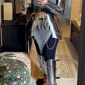 Puede incluir: Un conjunto de armadura de metal plateado con detalles de malla. La armadura incluye una pieza de pecho, hombreras, una falda y protectores de piernas. La armadura se lleva sobre unos pantalones negros.