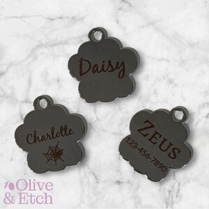 May include: Three silver paw-shaped pet ID tags with engraved names and phone numbers. The tags read "Daisy", "Charlotte" with a spider web design, and "ZEUS 123-456-7890".