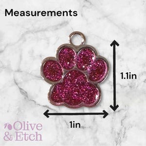 May include: Pink glitter paw print charm with a silver metal ring. The charm measures 1 inch wide and 1.1 inches tall.