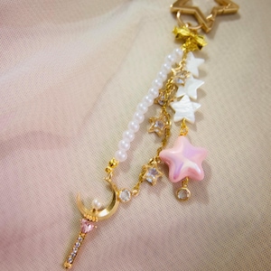 May include: A gold-colored star-shaped keychain with a dangling charm. The charm features a pearl strand, a crescent moon, a pink star, and smaller star and crystal accents. The background is a soft, neutral color.