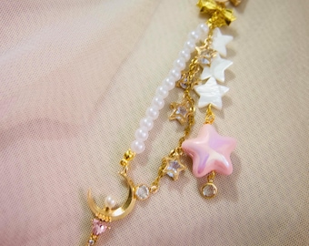 May include: A gold-colored star-shaped keychain with a dangling charm. The charm features a pearl strand, a crescent moon, a pink star, and smaller star and crystal accents. The background is a soft, neutral color.