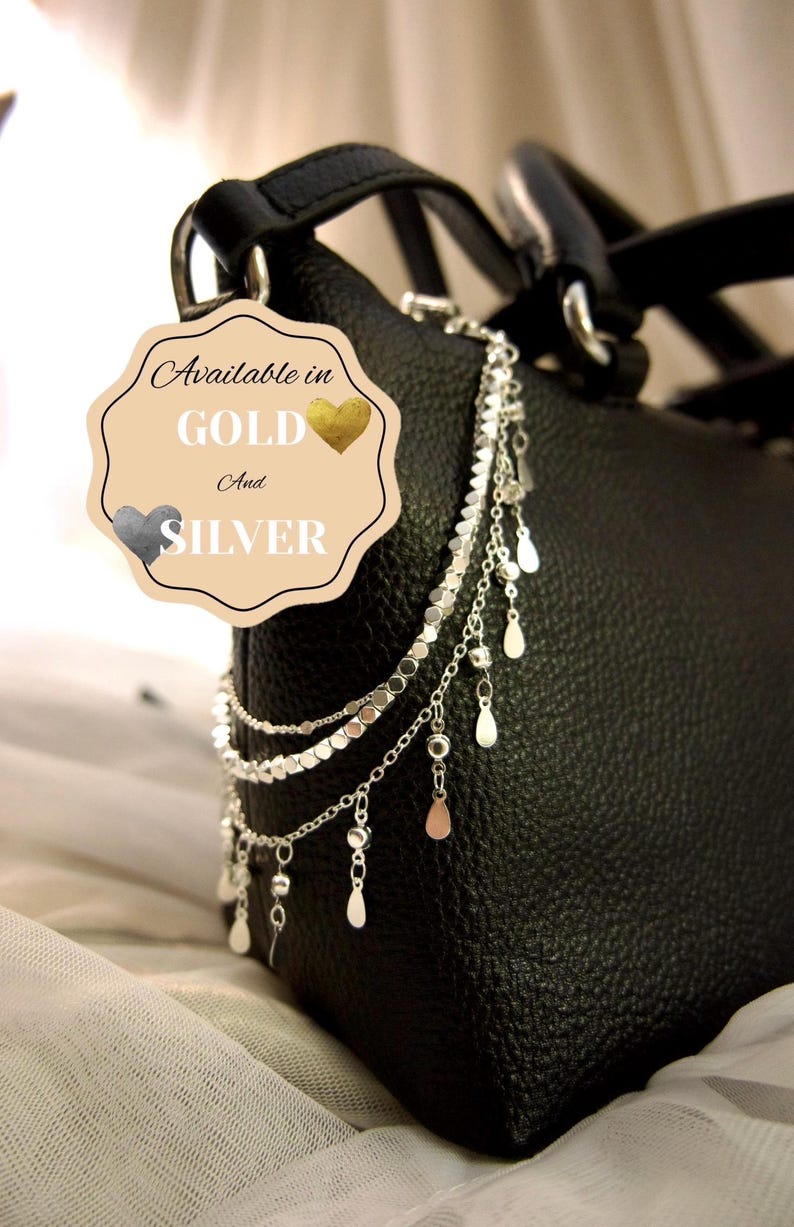 May include: A black textured handbag adorned with a silver-toned multi-strand chain accessory featuring small circular discs and dangling teardrop charms. A beige tag is attached, displaying the text: Available in GOLD And SILVER.