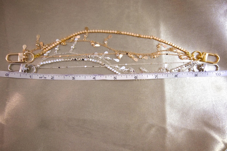 May include: Two decorative chain straps with lobster claw clasps at each end. One strap is gold-toned with dangling teardrop charms, and the other is silver-toned with flat circular discs. A white measuring tape shows a length of 10 inches.