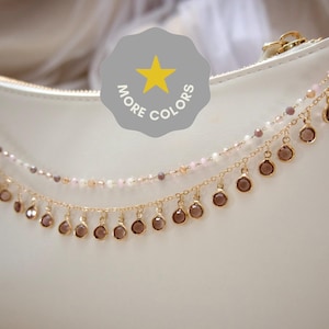 May include: A white handbag adorned with a double-layered gold-tone chain accessory. The top chain features small light pink beads, while the bottom chain displays a series of dangling round brown charms. A gray badge with a yellow star and the text MORE COLORS is visible.