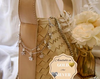 May include: A beige woven handbag with a solid top handle and a silver chain strap. The bag is decorated with a silver-toned charm chain featuring star and crystal accents. A label on the bag reads: Available in GOLD And SILVER.