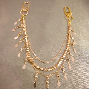 May include: A gold-toned multi-strand chain accessory featuring two lobster claw clasps at the ends. The design includes three layered chains adorned with small, flat, metallic teardrop-shaped charms and faceted gold beads hanging along the length.