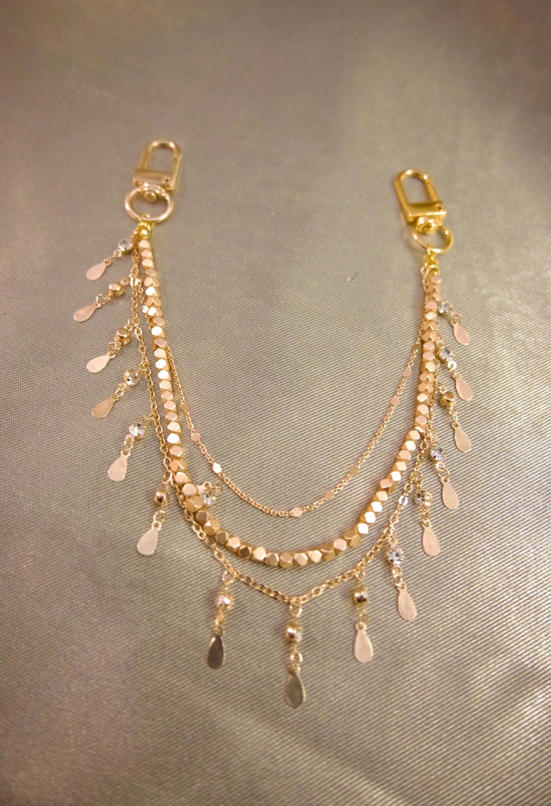 May include: A gold-toned multi-strand chain accessory featuring two lobster claw clasps at the ends. The design includes three layered chains adorned with small, flat, metallic teardrop-shaped charms and faceted gold beads hanging along the length.