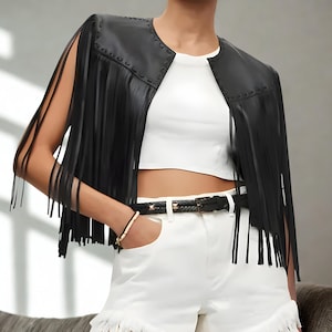K&ouml;nnte beinhalten: Schwarze Lederfransenjacke mit rundem Ausschnitt und offener Front. Die Jacke hat lange Fransen an den &Auml;rmeln und am Saum. Ein schwarzer G&uuml;rtel mit silberner Schnalle und wei&szlig;e Shorts vervollst&auml;ndigen den Look.