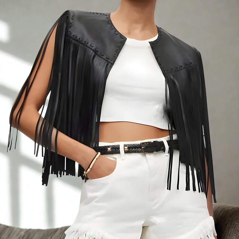 K&ouml;nnte beinhalten: Schwarze Lederfransenjacke mit rundem Ausschnitt und offener Front. Die Jacke hat lange Fransen an den &Auml;rmeln und am Saum. Ein schwarzer G&uuml;rtel mit silberner Schnalle und wei&szlig;e Shorts vervollst&auml;ndigen den Look.