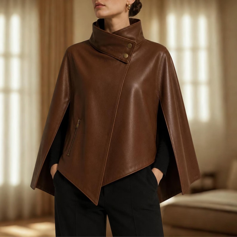 May include: A brown leather cape featuring a high collar with two gold-tone buttons, an asymmetrical front closure, and a vertical zippered pocket on the right side. The garment is worn over a black long-sleeved top and black trousers.