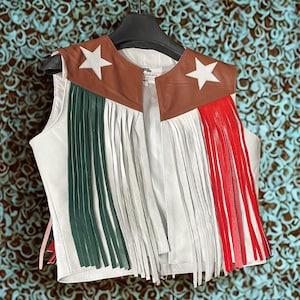 May include: White leather vest with fringe and brown leather accents. The vest has two stars on the front and the fringe is in green, white, and red.