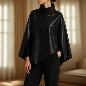 May include: A black faux leather cape featuring a high collar with two gold-toned buttons, an asymmetrical front closure, and a single vertical zippered pocket on the left side. The garment has wide arm openings and a structured, flared silhouette.