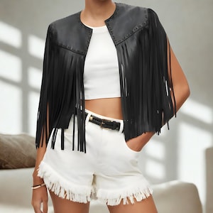 K&ouml;nnte beinhalten: Eine schwarze Kunstleder-Fransenweste mit Nieten. Die &auml;rmellose Weste hat lange Fransen. Das Model tr&auml;gt ein wei&szlig;es Crop-Top und wei&szlig;e Shorts mit Fransen.