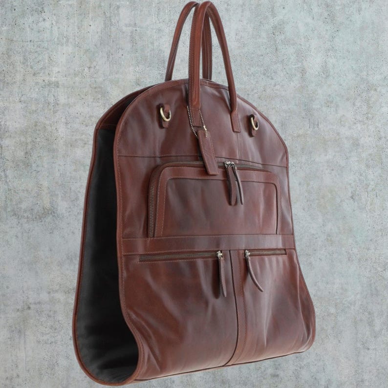K&ouml;nnte beinhalten: Eine braune Leder-Kleidertasche mit mehreren Taschen und einem Rei&szlig;verschluss. Die Tasche hat einen Griff und einen Kleiderb&uuml;gel.