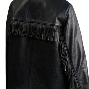 May include: Black leather jacket with fringe detail across the upper back and down the sleeves. The jacket has a classic design with a textured finish, offering a stylish and edgy look. The fringe adds a unique, decorative element.