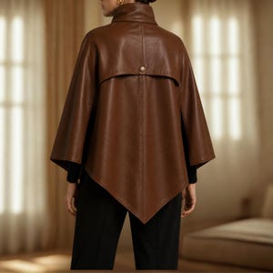 May include: A brown leather cape featuring a high collar, a pointed hemline, and a single gold-toned button detail at the upper back. The garment is worn over a black long-sleeved top and black trousers, showcasing a structured, sleeveless design.