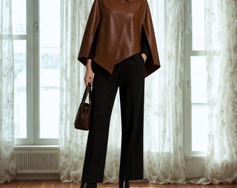 May include: A brown leather cape with a high collar and a single button closure, worn over black trousers and a black long-sleeved top. The outfit is paired with black leather ankle boots and a matching brown leather handbag held in one hand.
