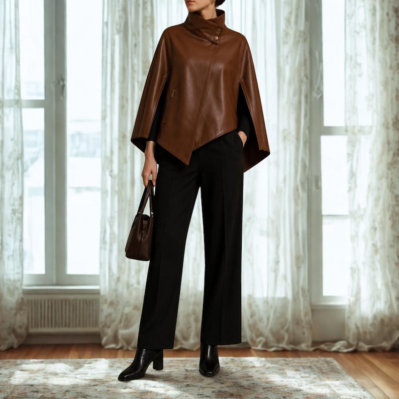 May include: A brown leather cape with a high collar and a single button closure, worn over black trousers and a black long-sleeved top. The outfit is paired with black leather ankle boots and a matching brown leather handbag held in one hand.