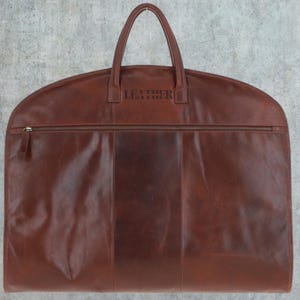 K&ouml;nnte beinhalten: Eine braune Leder-Kleidertasche mit Rei&szlig;verschluss und Tragegriff. Auf der Vorderseite ist das Wort "LEATHER" gepr&auml;gt.