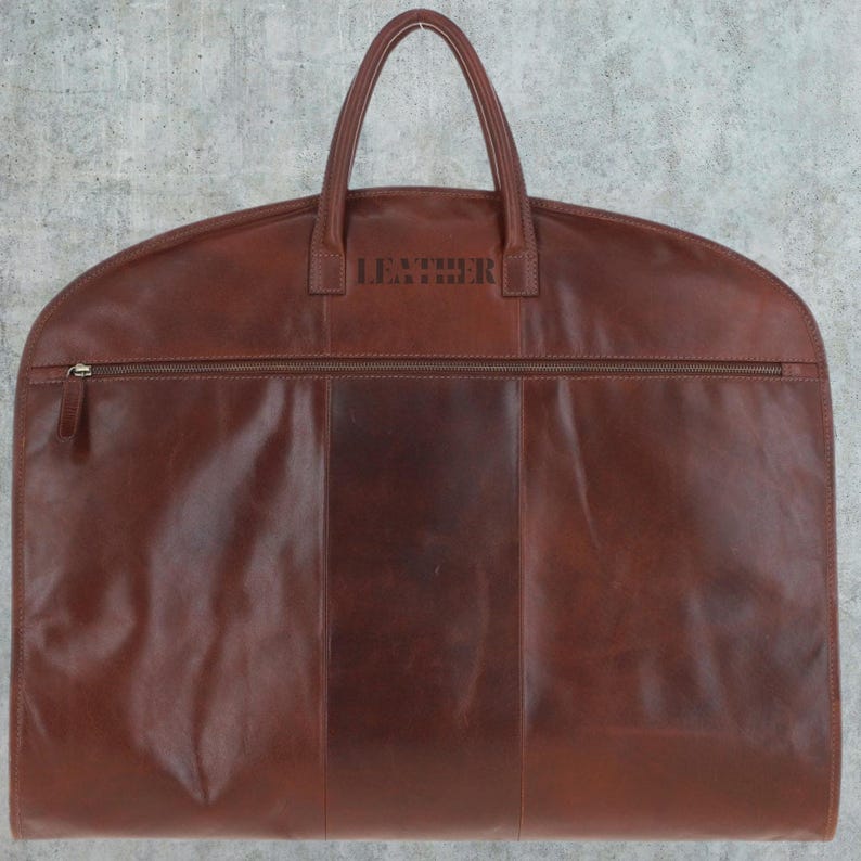 K&ouml;nnte beinhalten: Eine braune Leder-Kleidertasche mit Rei&szlig;verschluss und Tragegriff. Auf der Vorderseite ist das Wort "LEATHER" gepr&auml;gt.