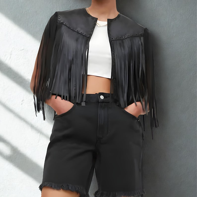 K&ouml;nnte beinhalten: Schwarze Lederjacke mit Fransen und kurzem Schnitt. Die Jacke hat einen runden Ausschnitt und wird mit schwarzen Shorts kombiniert. Die Shorts haben einen ausgefransten Saum und eine hohe Taille. Das Outfit ist eine moderne Interpretation eines klassischen Stils.