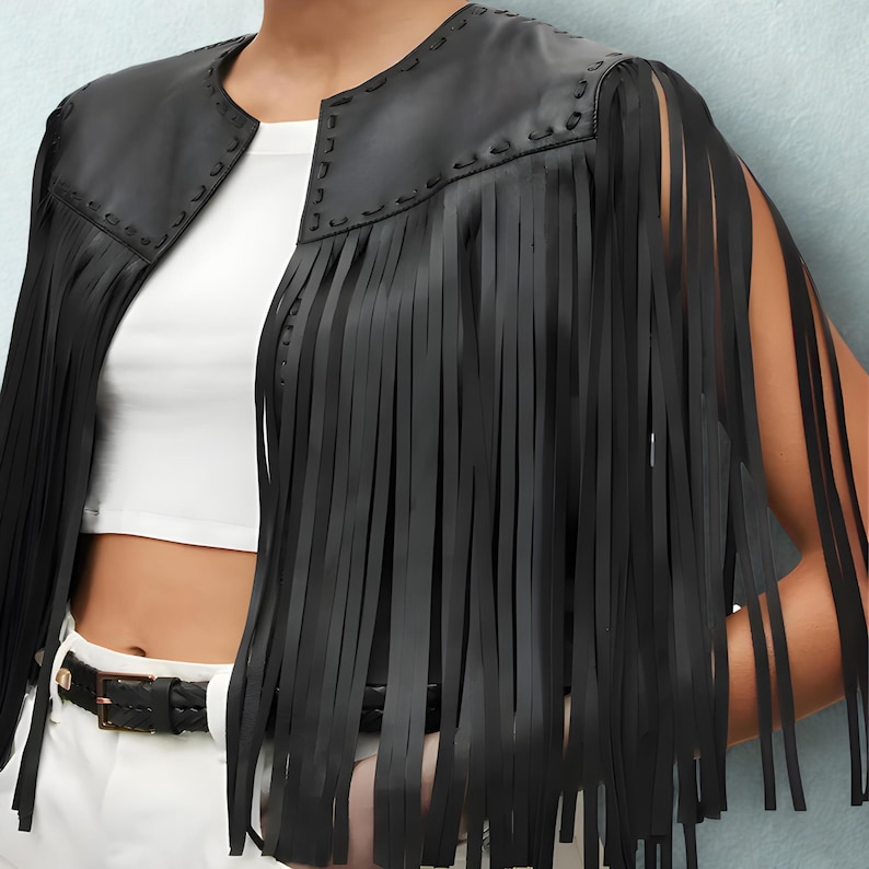 K&ouml;nnte beinhalten: Schwarze Lederjacke mit langen Fransen. Die Jacke hat einen runden Ausschnitt und offene &Auml;rmel. Die Fransen h&auml;ngen von den Schultern und um die Arme. Die Jacke wird &uuml;ber einem wei&szlig;en Oberteil getragen und mit einem schwarzen G&uuml;rtel kombiniert.