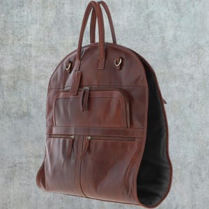 K&ouml;nnte beinhalten: Eine braune Leder-Kleidertasche mit mehreren Taschen und einem Rei&szlig;verschluss. Die Tasche hat eine abgerundete Oberseite und zwei Griffe.