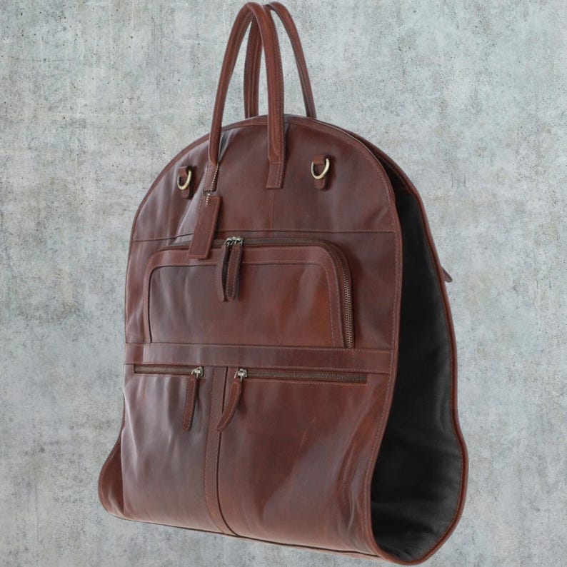 K&ouml;nnte beinhalten: Eine braune Leder-Kleidertasche mit mehreren Taschen und einem Rei&szlig;verschluss. Die Tasche hat eine abgerundete Oberseite und zwei Griffe.