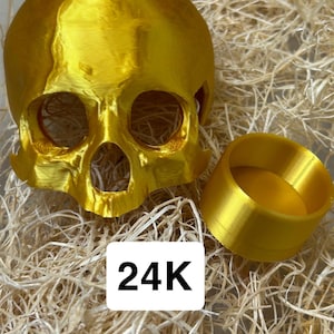 May include: A gold-colored skull sculpture and a matching ring-shaped object. The skull has visible eye sockets and nasal cavity. The objects are on a bed of straw-like material. The text "24K" is displayed on a white rectangle.