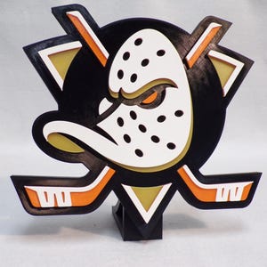 May include: A 3D-printed logo of the Anaheim Ducks hockey team. The logo features a duck-masked goalie with a black background, orange and white accents, and crossed hockey sticks. The logo is mounted on a black base.