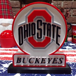 May include: A lighted Ohio State Buckeyes sign with a red and white logo and the words "OHIO STATE" in black. The base reads "BUCKEYES". The sign is on a table with a red, white, and blue patterned tablecloth.