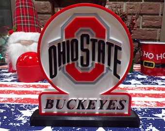 May include: A lighted Ohio State Buckeyes sign with a red and white logo and the words "OHIO STATE" in black. The base reads "BUCKEYES". The sign is on a table with a red, white, and blue patterned tablecloth.