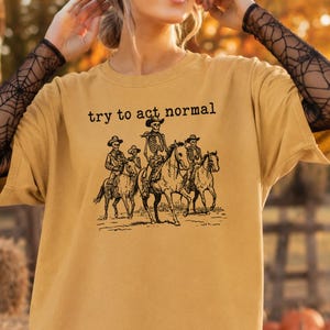 May include: A mustard yellow short-sleeve t-shirt featuring a black graphic print of four skeletons wearing cowboy hats and riding horses. The text "try to act normal" is printed in black above the graphic. The shirt has a crew neckline.