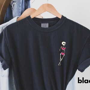 May include: Black t-shirt featuring a small skeleton graphic wearing a pink bikini. The skeleton is drawn in a cartoon style. The word "black" is printed in white on the lower right side of the shirt.