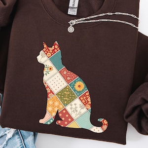 May include: Dark chocolate-colored sweatshirt featuring a patchwork cat design. The cat silhouette is composed of various patterned squares in shades of red, yellow, green, and blue. A silver necklace with a pendant is draped across the sweatshirt.