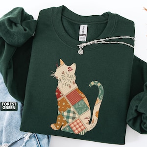 以下が含まれることがあります： パッチワークの猫のデザインが施されたフォレストグリーンのスウェットシャツ。猫のシルエットは、赤、オレンジ、緑、茶色の花柄と幾何学模様で埋められています。小さなペンダントが付いたシルバーのネックレスがスウェットシャツにかけられています。