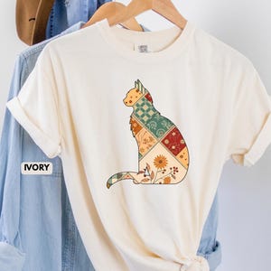 May include: Ivory-colored t-shirt featuring a patchwork cat design. The cat silhouette is filled with various patterns, including floral and geometric designs in shades of orange, green, and red. The shirt is hanging on a wooden hanger.