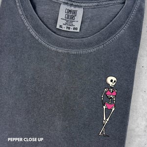 May include: A charcoal gray t-shirt with a crew neckline and a Comfort Colors brand label. The shirt features a small graphic of a skeleton wearing a pink bikini. The text PEPPER CLOSE UP is visible in the bottom left corner of the image.